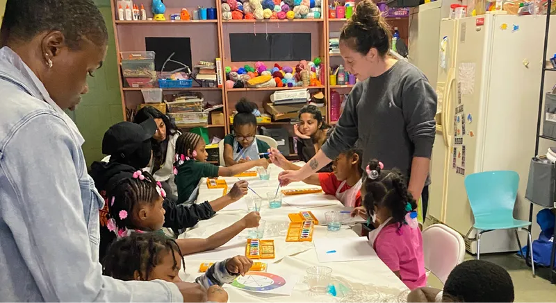 Many small children sitting around a long table with paints of various colors.